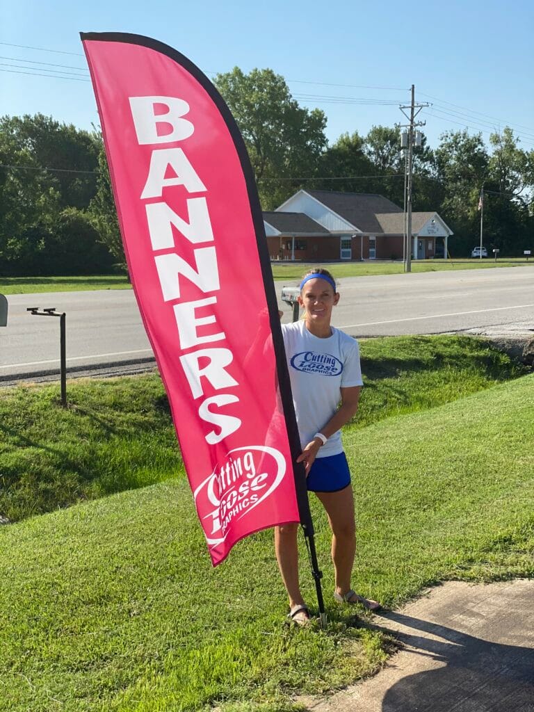 Clarissay Hallacy of Cutting Loose Graphics standing next to a giant red Flag Banner by the road on North Main in Joplin, MO.