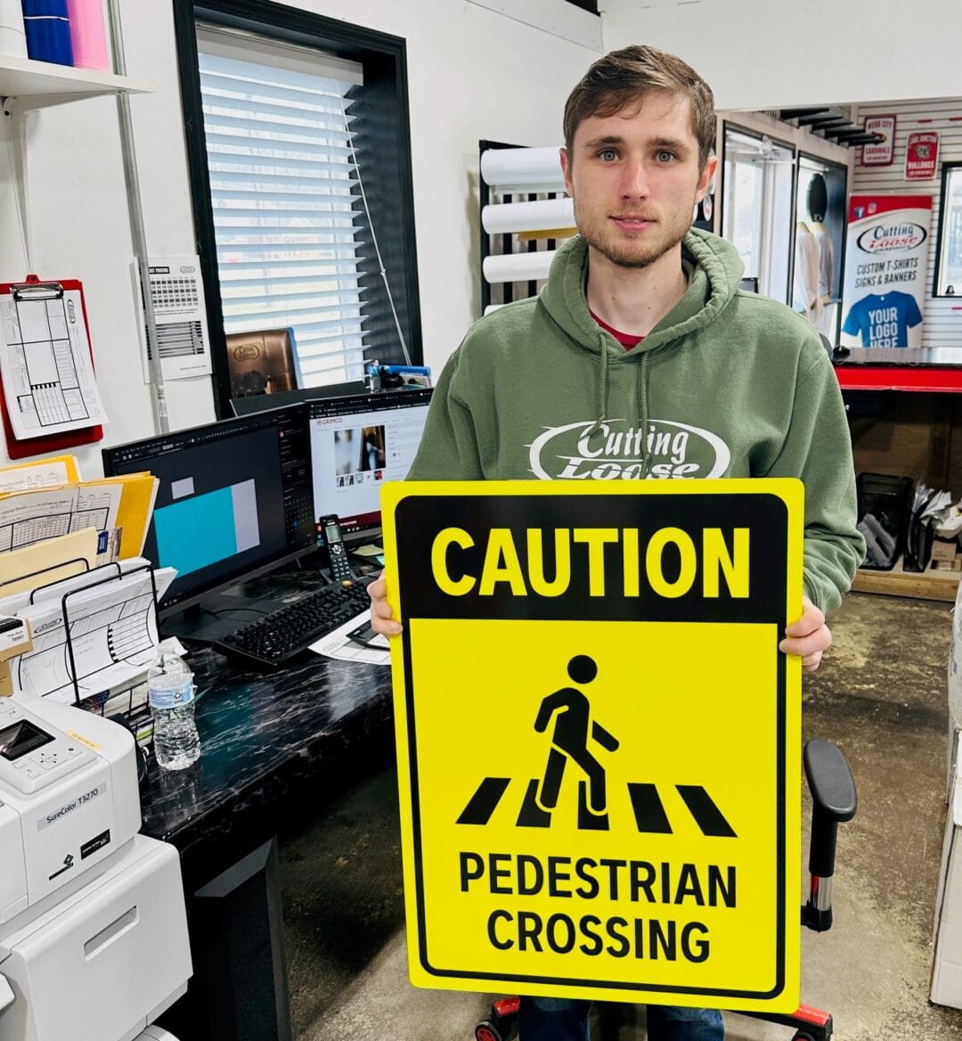 Cole of Cutting Loose Graphics holding a yellow 'Caution Pedestrian Crossing" sign.